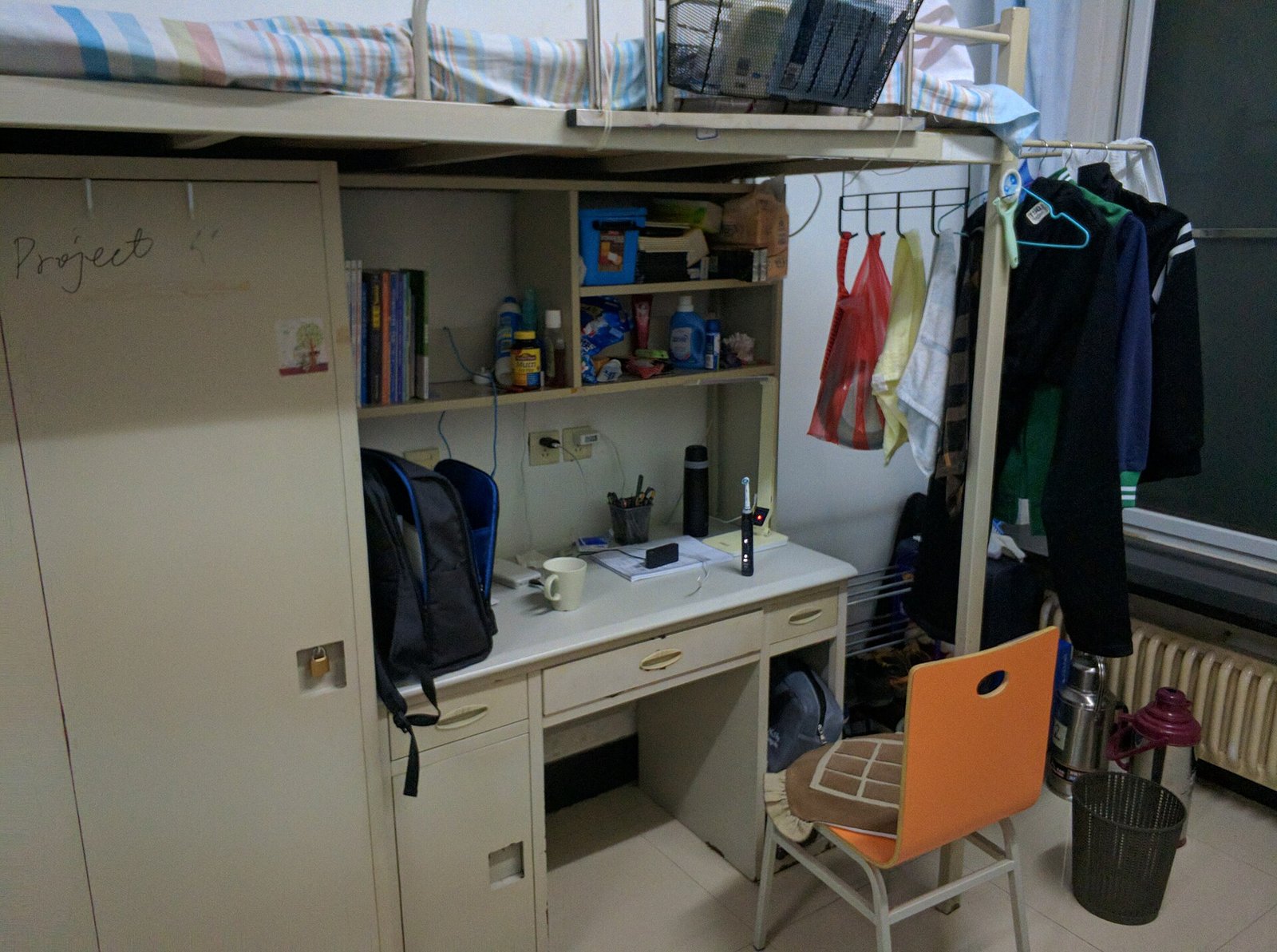 A college dorm room with a loft bed above a desk, bookshelf and backpack.