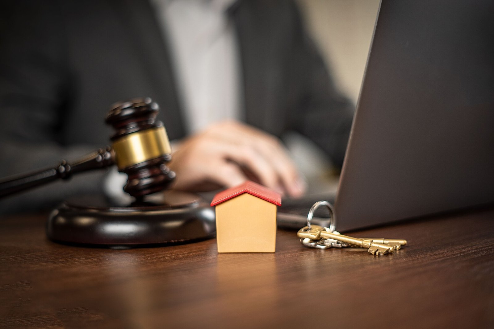 A gavel, a small model house and keys on a desk beside a laptop.