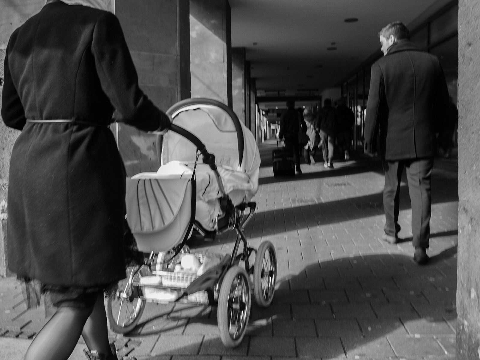A parent pushes a baby stroller along a city walkway, seen from behind.