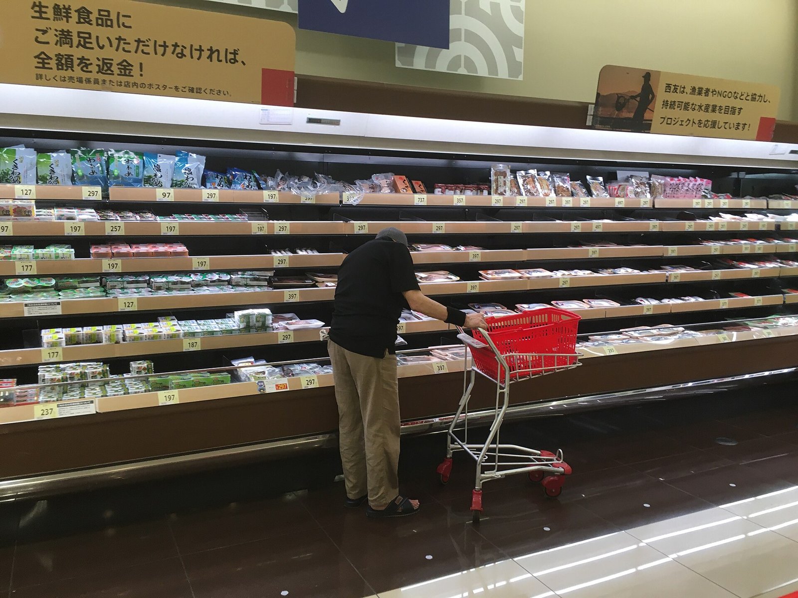 A shopper with a red cart browses a long supermarket display case, seen from behind.