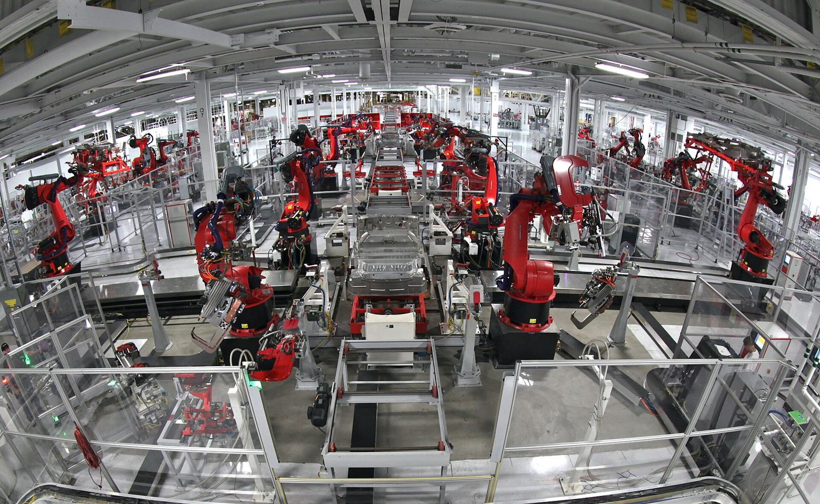 Red robotic arms work on car bodies along a factory assembly line.