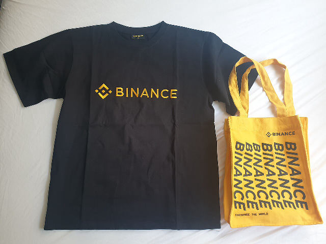 A black Binance-branded T-shirt with the Binance logo next to a yellow Binance tote bag